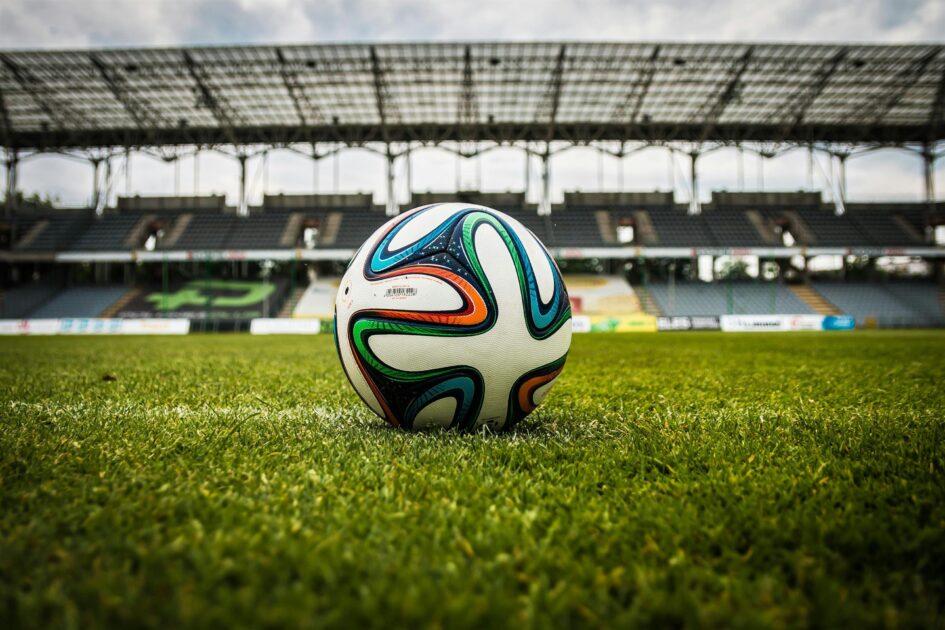 En fotboll ligger på en grön gräsplan i en tom fotbollsarena med tak och läktare i bakgrunden.