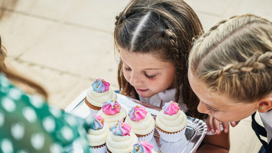 Two schoolgirls admiring delicious cupcakes held by teacher