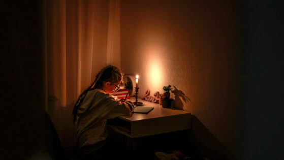 Schoolgirl uses candlelight to writes and reads in darkness without electric lights at home. Teen with poor eyesight wears glasses to studies and to do school homework during blackout. Vision problems